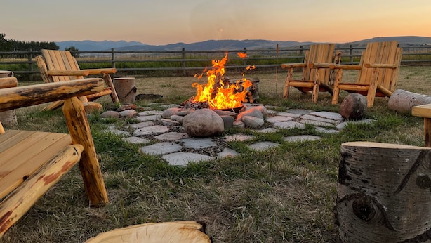 A campfire with wooden chairs arranged around it, set in a grassy area with a fence and mountains in the background. - Home w/river access & stunning views of the mountains in Gallatin Gateway - Le Ciel (Gallatin Gateway)
