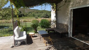Een terras buiten met een witte fauteuil met kussens, een houten bank en een kleine ronde tafel.