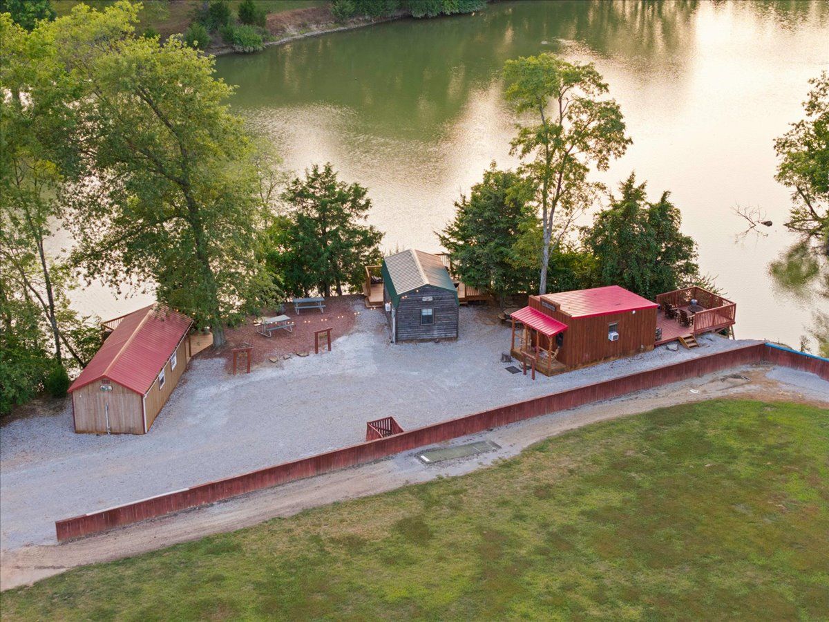 A lakeside property with a gravel patio, a wooden cabin, a red-roofed structure, and a fenced area.