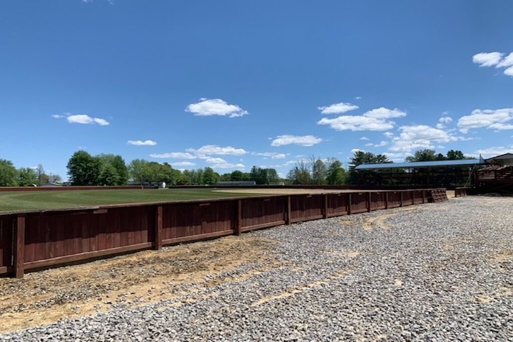 A gravel sports field with a wooden fence and a covered seating area.