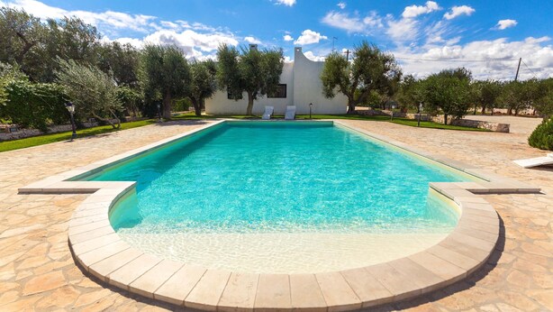 Una piscina rectangular con solárium de piedra, rodeada de árboles y un edificio blanco con tejado inclinado.