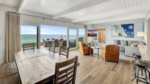 A spacious living area with a wooden floor, a dining table, and a sofa. Large windows offer a view of the ocean. - ON THE SAND!! Private Beach! OCEAN FRONT HOT TUB!! STR14-0126 (Dana Point)