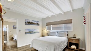 A bedroom with a large bed, wooden flooring, white walls, and a ceiling with exposed beams. There are two paintings on the wall and a nightstand with a lamp. - ON THE SAND!! Private Beach! OCEAN FRONT HOT TUB!! STR14-0126 (Dana Point)