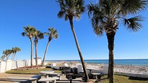 A patio area with stone tables and benches, palm trees, and a view of the beach and ocean. - El Matador 316 by AJH Rentals FREE BEACH SERVICE+ACTIVITIES! $300/Day (Fort Walton Beach)