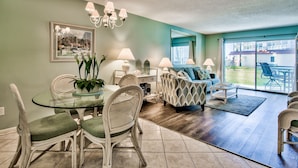 A spacious living area with a dining table, chairs, a sofa, and a coffee table. There is a large window providing a view of the outdoors. - El Matador 316 by AJH Rentals FREE BEACH SERVICE+ACTIVITIES! $300/Day (Fort Walton Beach)