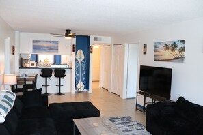 A living room with a sofa, a coffee table, and a TV. There is a kitchen area with a microwave and a surfboard decoration. - Emerald Isle 404 by Alicia Hollis Rentals FREE TICKETS! $300/Day (Fort Walton Beach)