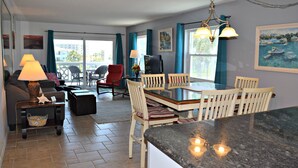 A spacious living area with a dining table, chairs, and a kitchen island. There is a sofa, a TV, and a lamp in the living area. The room has large windows with curtains.