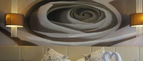 Une chambre d’hôtel avec un grand tableau abstrait au mur, un lit recouvert de draps blancs, deux coussins à motifs floraux et un cygne décoratif fait de serviettes posé sur le lit.