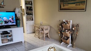 Una sala de estar con televisor, una mesa de centro de vidrio con objetos decorativos, una silla de mimbre, una estantería y un cuadro en la pared.