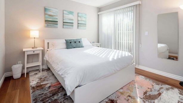A bedroom with a white bed, a bedside table, a lamp, and a large window with blinds. - Bright, Quiet, Comfy 1BR Cottage in Mountain View (Mountain View)