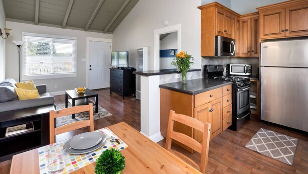 A modern kitchen with wooden cabinets, a stainless steel refrigerator, and a microwave. There is a dining area with a table set for two and a sofa in the living room. - Spacious, Soaring Ceilings, Near Downtown MV, GOOG (Mountain View)