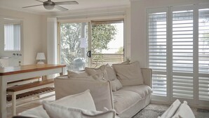 Ein Wohnzimmer mit einer Couch, einem Holztisch, einem Deckenventilator und großen Fenstern mit Jalousien.