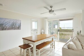 Habitación con una mesa de madera, banco y ventilador de techo. Posee un ventanal corredero que da a un espacio exterior con vista al mar.