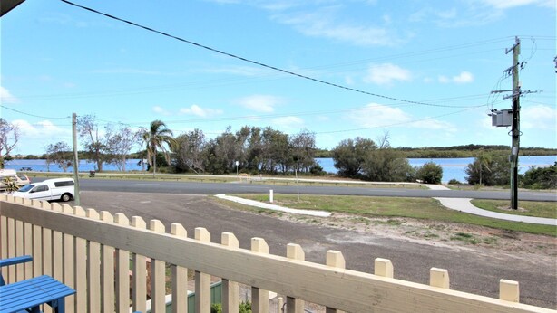A view from a balcony overlooking a road with a white car parked on the side. There is a body of water in the background, and the area is surrounded by trees and a fence. - Bear's a well located fisherman and family holiday home in Harrington (Harrington)