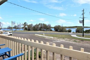 A view from a balcony overlooking a road with a white car parked on the side. There is a body of water in the background, and the area is surrounded by trees and a fence. - Bear's a well located fisherman and family holiday home in Harrington (Harrington)