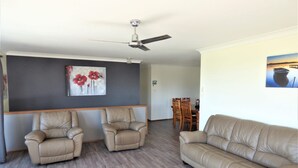 A living room with a sofa, two armchairs, a dining table with chairs, a ceiling fan, and a painting on the wall. - Bear's a well located fisherman and family holiday home in Harrington (Harrington)