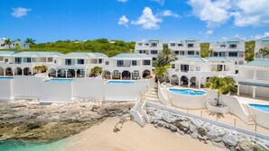 Un complexe hôtelier composé de plusieurs bâtiments blancs, chacun doté d’une piscine, qui se déploie sur fond de ciel bleu azur et de plage rocheuse.