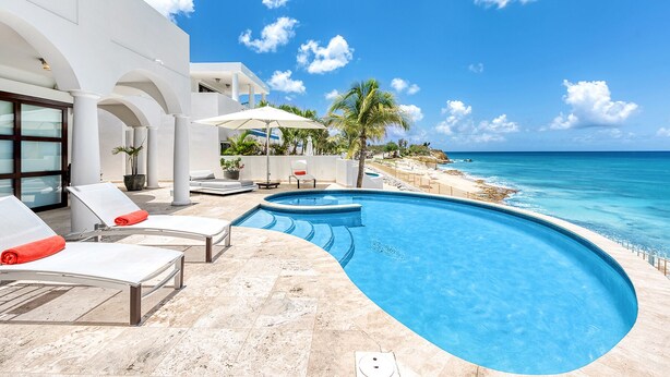 Un espace piscine moderne, doté de chaises longues et de parasols, avec vue sur une plage et l’océan.