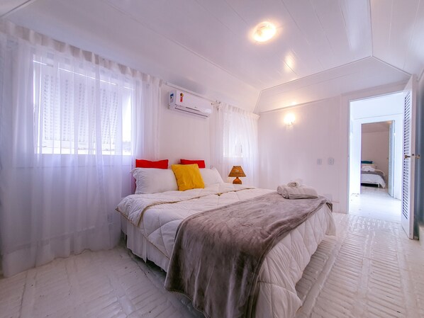 Un dormitorio con cama, cortinas blancas, una lámpara y un baño visible a través de una puerta abierta.