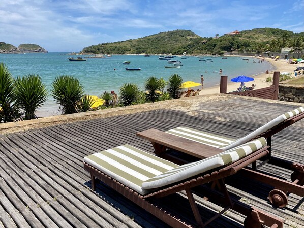 Una terraza de madera con sillones de descanso y una mesa, con vista a una playa con barcos y gente.