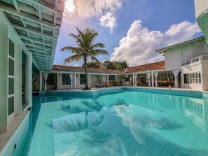 A swimming pool with tiled flooring, a palm tree, and a covered outdoor area with seating.