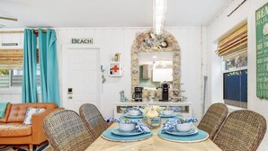 A dining area with a wooden table set for four, surrounded by wicker chairs. A mirror with a decorative frame is above the table, and a sign on the wall reads "BEACH".