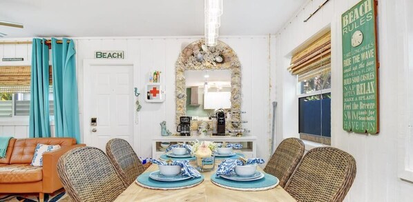 A dining area with a wooden table set for four, surrounded by wicker chairs. A mirror with a decorative frame is above the table, and a sign on the wall reads "BEACH".