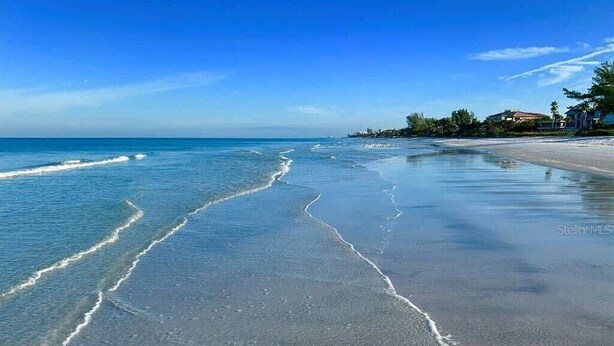 A beach with clear blue water and a sandy shore. There are houses in the background.