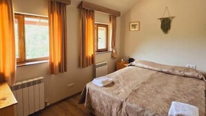 A bedroom with a large bed, two windows with curtains, a radiator, a bedside table, and a hanging plant. - Ecokayan Boutique Hotel & Camping (Dilijan)