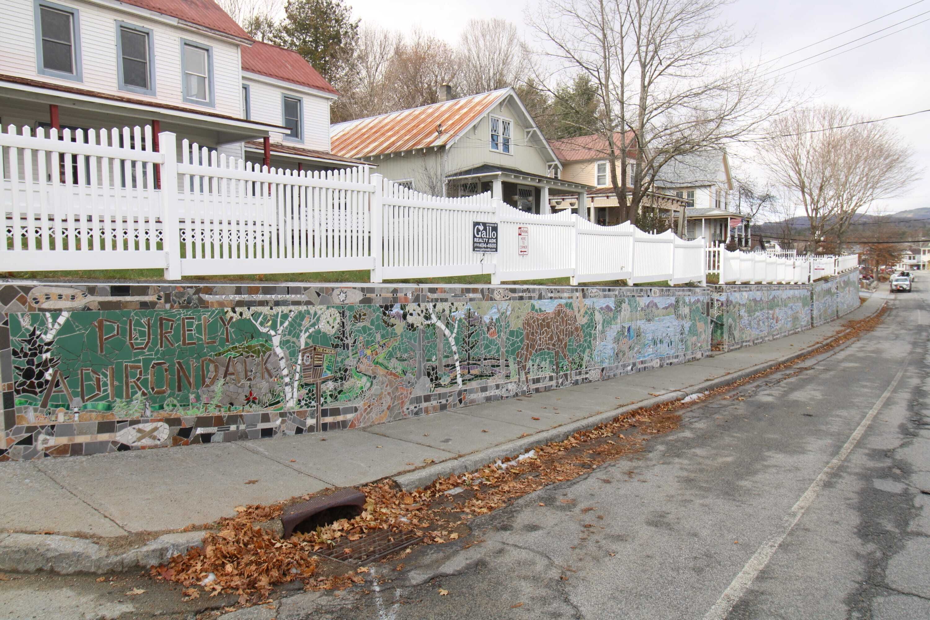 A residential street with houses, a white picket fence, and a mosaic wall with graffiti.