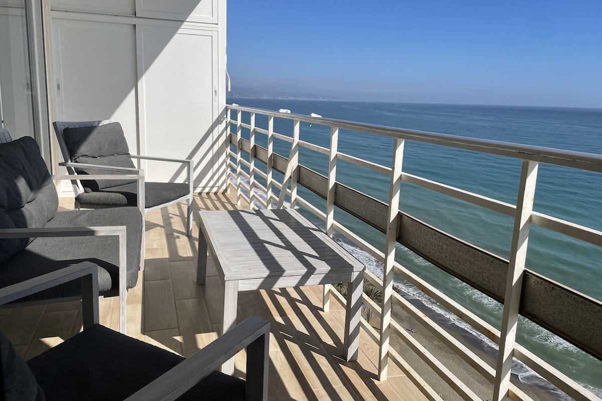 A balcony with a view of the ocean, featuring a railing, a table, and two chairs.
