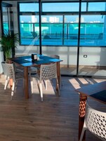 Un comedor con una mesa y sillas de madera, ventanas amplias con vista a un cuerpo de agua y edificios, y una planta en maceta.