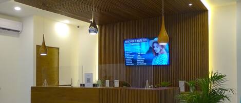 A modern reception desk with a wooden finish, a digital screen displaying information, and a potted plant.