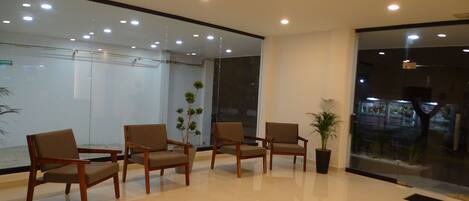A modern waiting area with wooden chairs and potted plants, a glass wall, and recessed ceiling lights.