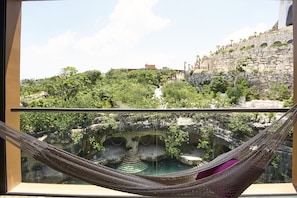 A hammock in a window overlooking a lush garden and a stone structure with a pool.