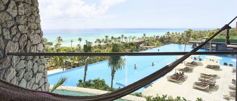 A hammock on a balcony overlooking a pool and beach area with palm trees and a view of the ocean.