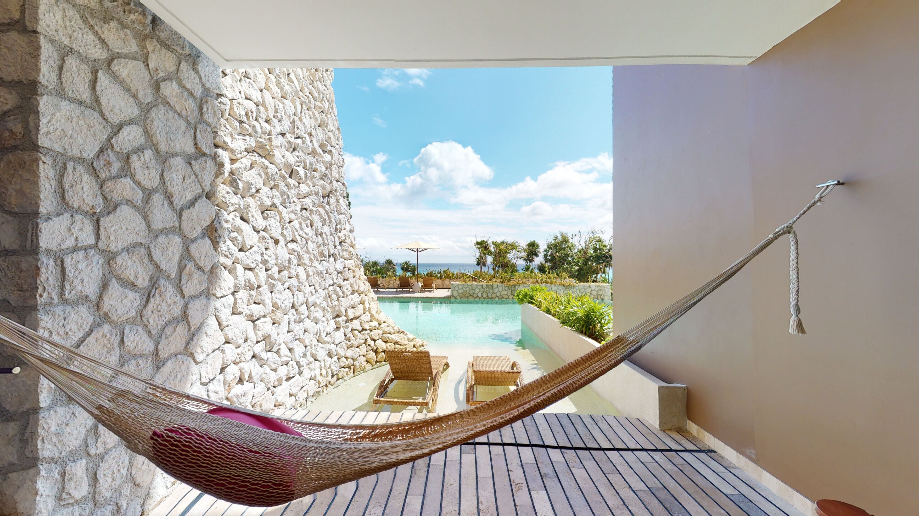 A hammock on a balcony overlooking a pool with stone walls and outdoor furniture.