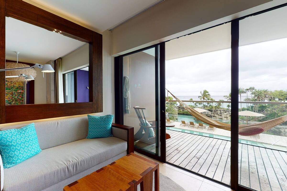 Una habitación moderna con un ventanal grande, un sofá con cojines estampados, una mesa de madera y una hamaca en un balcón exterior con vista a la piscina.