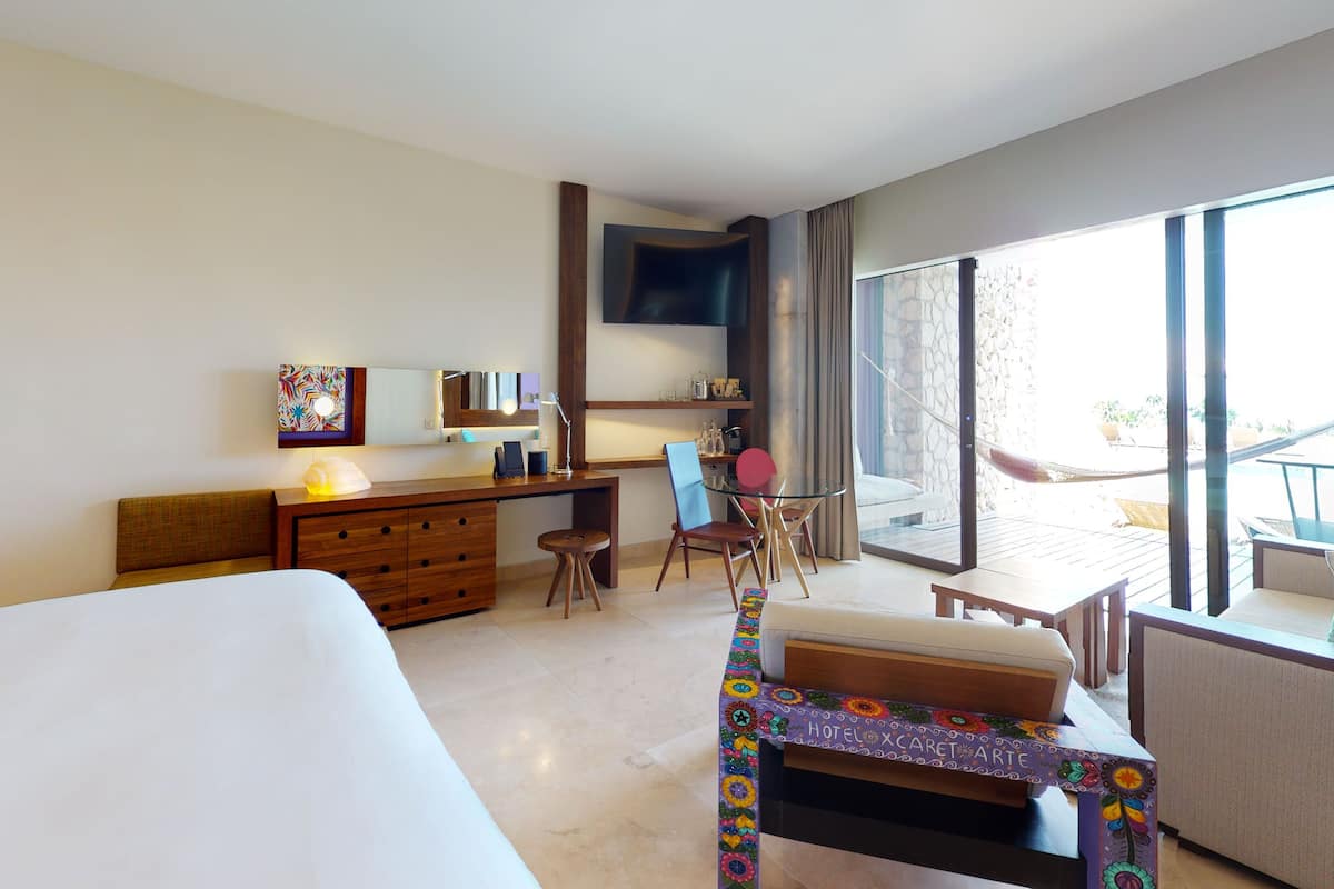 Habitación de hotel con una cama grande, un escritorio con una silla, una mesita y un balcón con hamaca.
