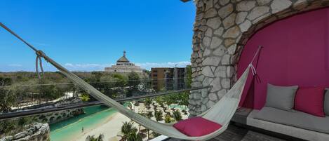 A hammock chair with a pink cushion on a balcony overlooking a beach and a building with a dome.