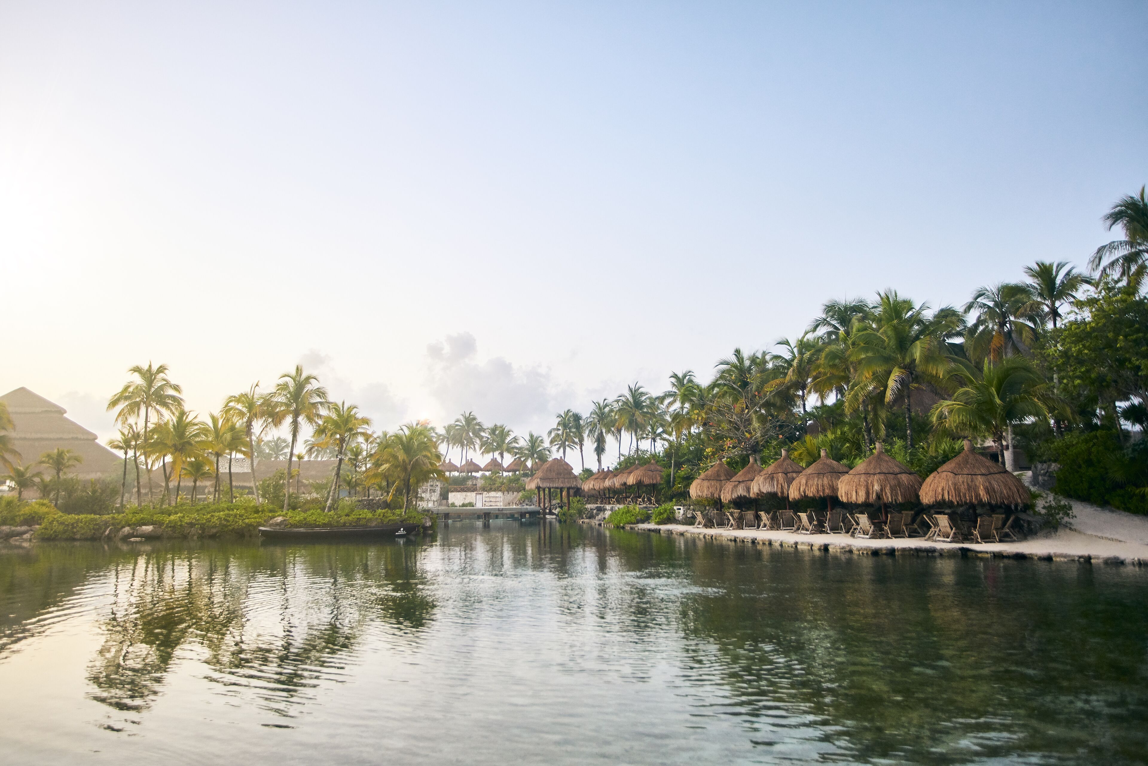 A serene resort pool area with thatched umbrellas and palm trees, surrounded by lush greenery and a clear sky.