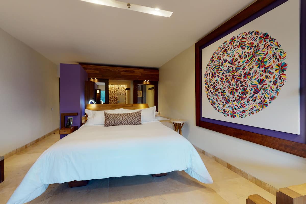 Una habitación de hotel moderna con una cama grande, una mesita de noche, un espejo y un cuadro abstracto colorido en la pared.