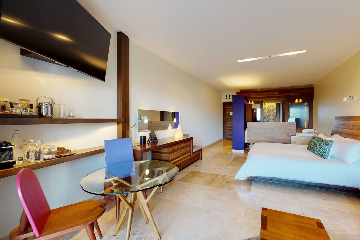 Habitación de hotel moderna con cama, escritorio, silla, televisión y baño visible a través de una pared divisoria.