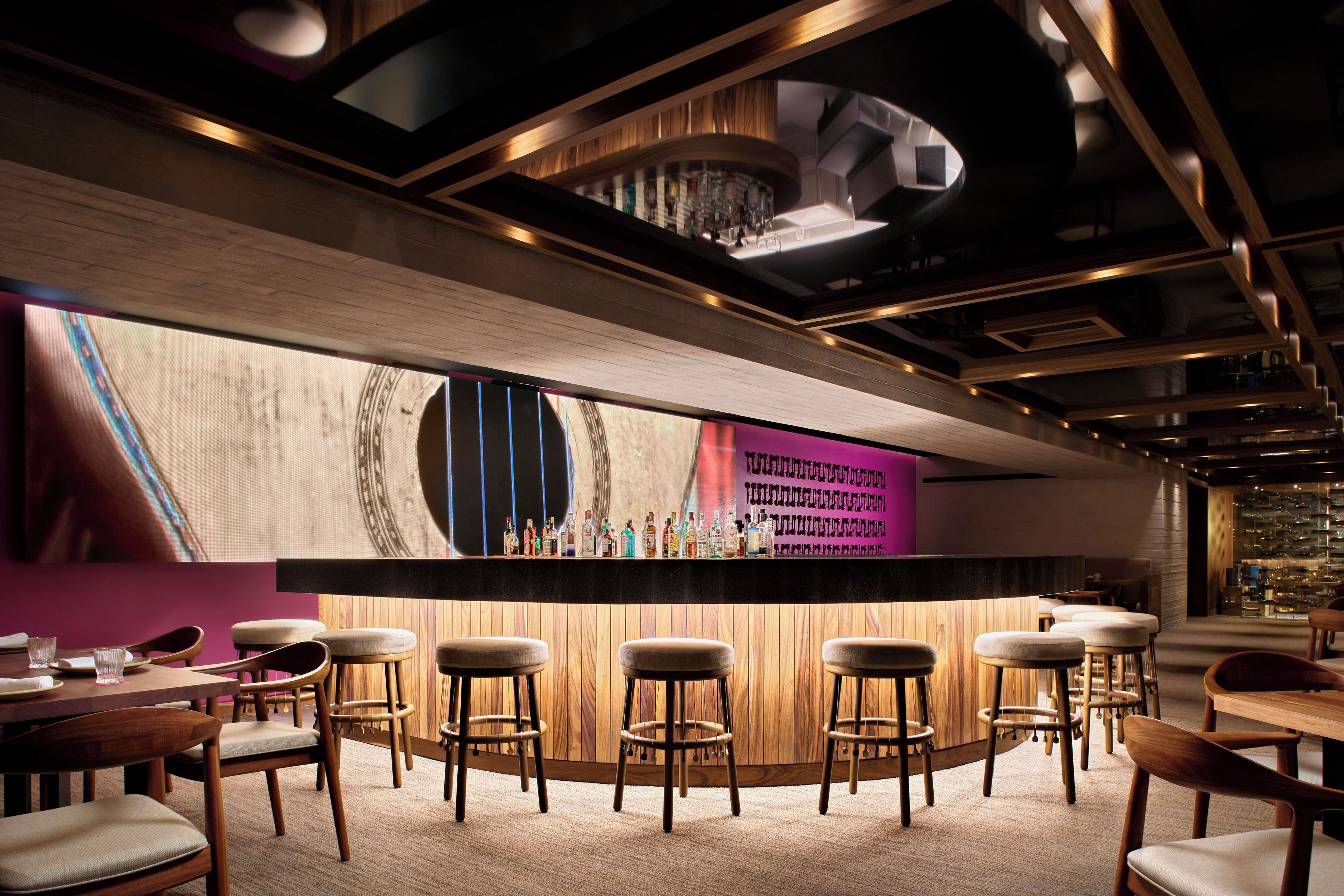 Zona de bar moderna con taburetes de madera, una barra con botellas y vista del área del bar a través de un ventanal grande.