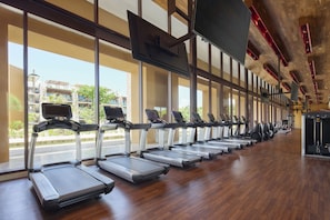 A gym with treadmills aligned along a wall with large windows, a wooden floor, and exercise equipment.