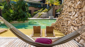 Una hamaca con una almohada roja sobre una terraza de madera junto a una piscina con dos sillas de mimbre.