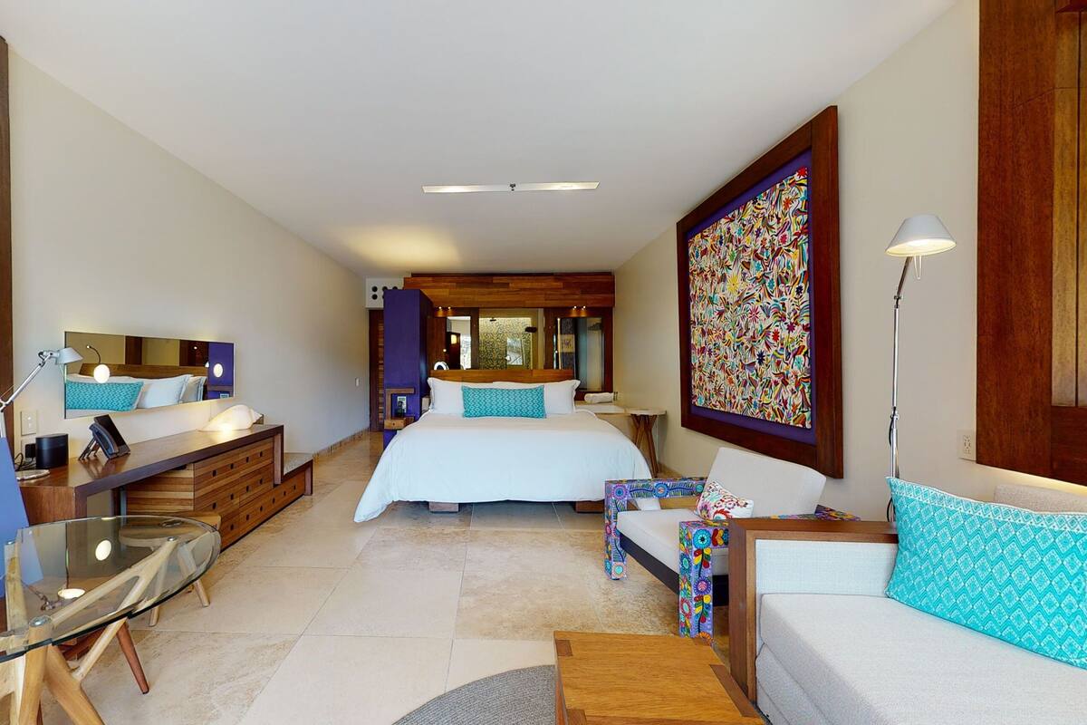 Una habitación de hotel moderna con una cama grande, un escritorio con una computadora, una mesa de vidrio y un ventanal amplio.