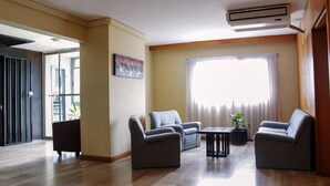 A spacious living area with a wooden floor, two grey sofas, a small table, and a large window with curtains. - Residence Les Hauts de L Indenie (Abidjan)