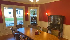 Une salle à manger avec une grande table en bois entourée de chaises, deux fenêtres donnant sur la rue et un buffet en bois foncé doté de portes en verre.
