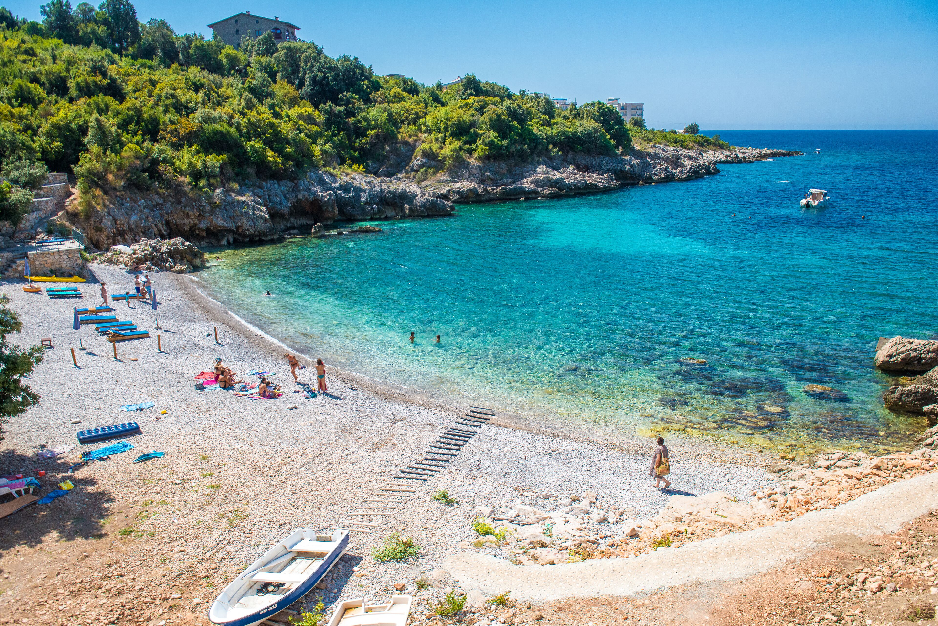 Una playa con agua turquesa cristalina, costa rocosa y algunos barcos.